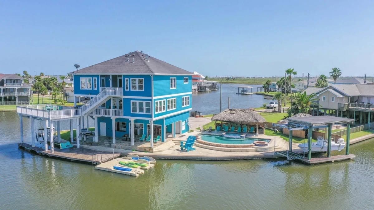 Aerial view of a three-story teal canal-front home in Jamaica Beach, Galveston with pool, palapa, and private boat dock.